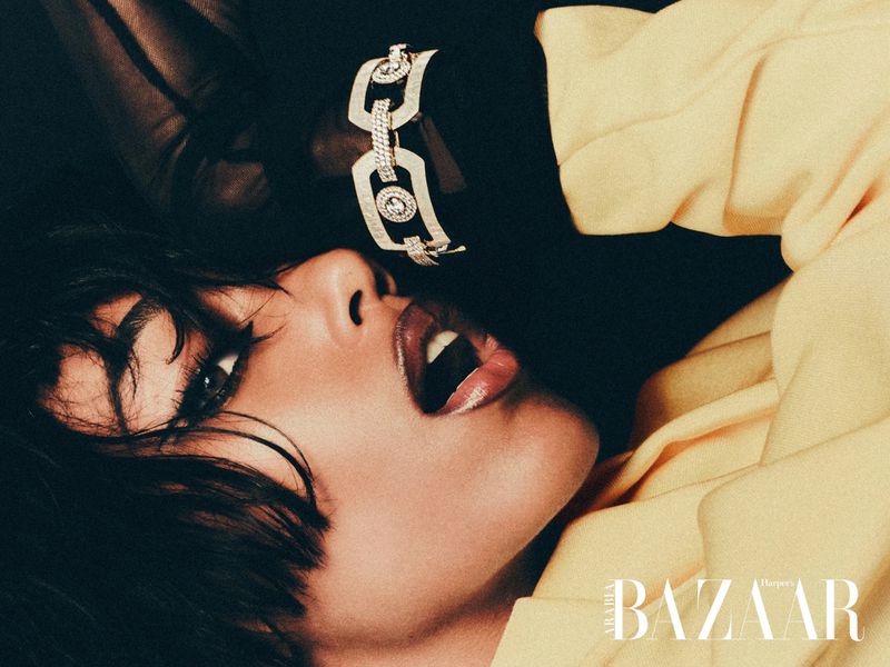Woman with open mouth wearing a yellow jacket and diamond bracelet, laying down in a dark studio; Bazaar magazine logo at the bottom.