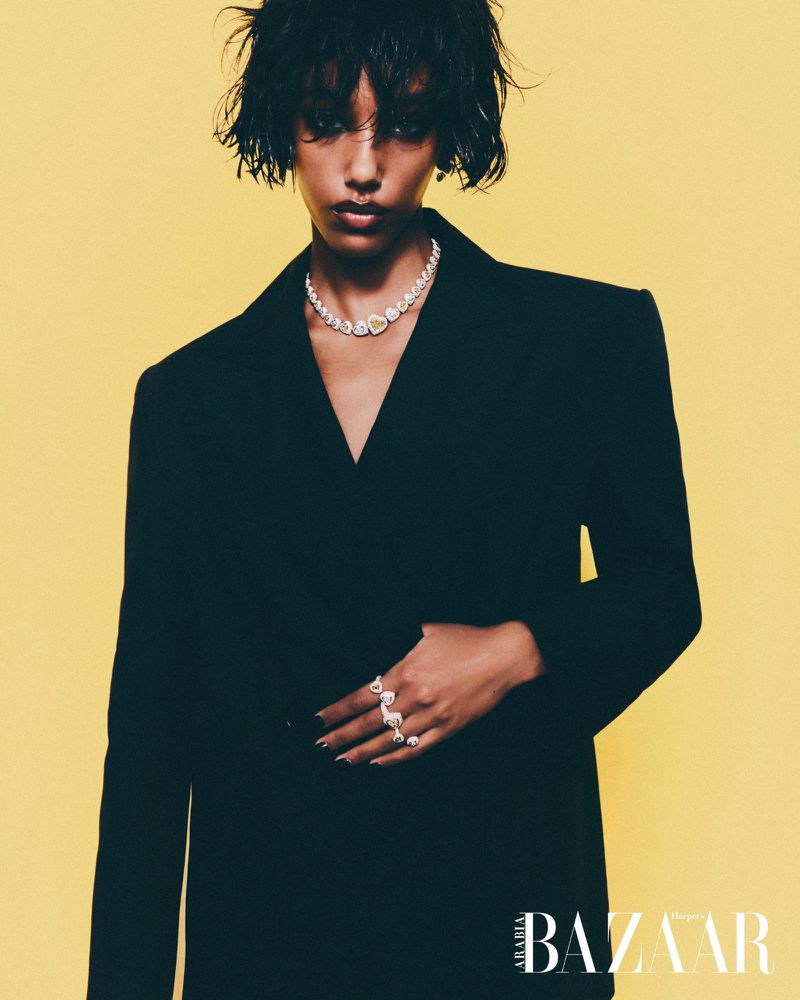 Woman in a black oversized blazer with a diamond necklace and rings poses in front of a solid yellow background in a fashion editorial.