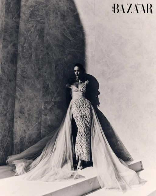 Fashion shot of a woman in a light-colored gown posing against a marble wall with natural lighting and a magazine logo. 
