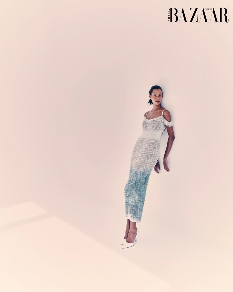 Elegant woman in shimmering gown poses against a soft pink backdrop, accentuated by a sleek hairstyle and matching pointed-toe high heels.