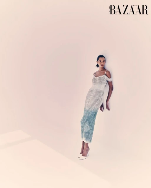 Elegant woman in shimmering gown poses against a soft pink backdrop, accentuated by a sleek hairstyle and matching pointed-toe high heels.