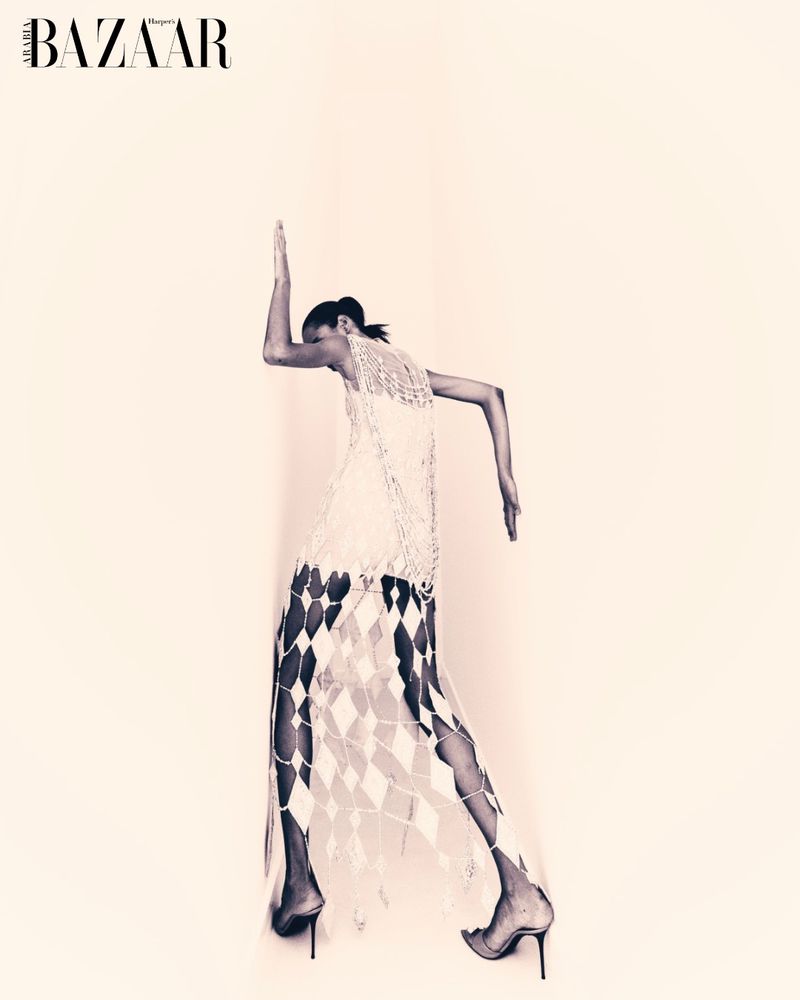 Monochrome fashion shot of a woman in a light-colored, sheer, diamond-patterned dress and high heels, posing against an off-white backdrop.