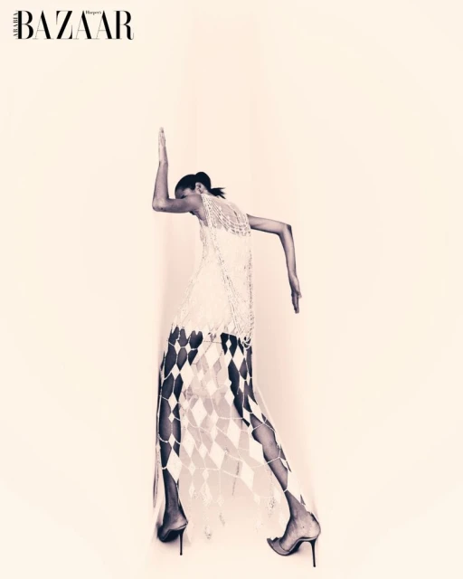 Monochrome fashion shot of a woman in a light-colored, sheer, diamond-patterned dress and high heels, posing against an off-white backdrop.