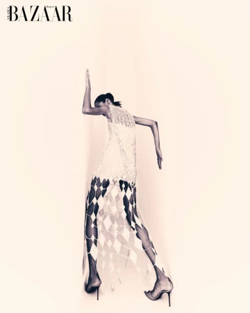Monochrome fashion shot of a woman in a light-colored, sheer, diamond-patterned dress and high heels, posing against an off-white backdrop.