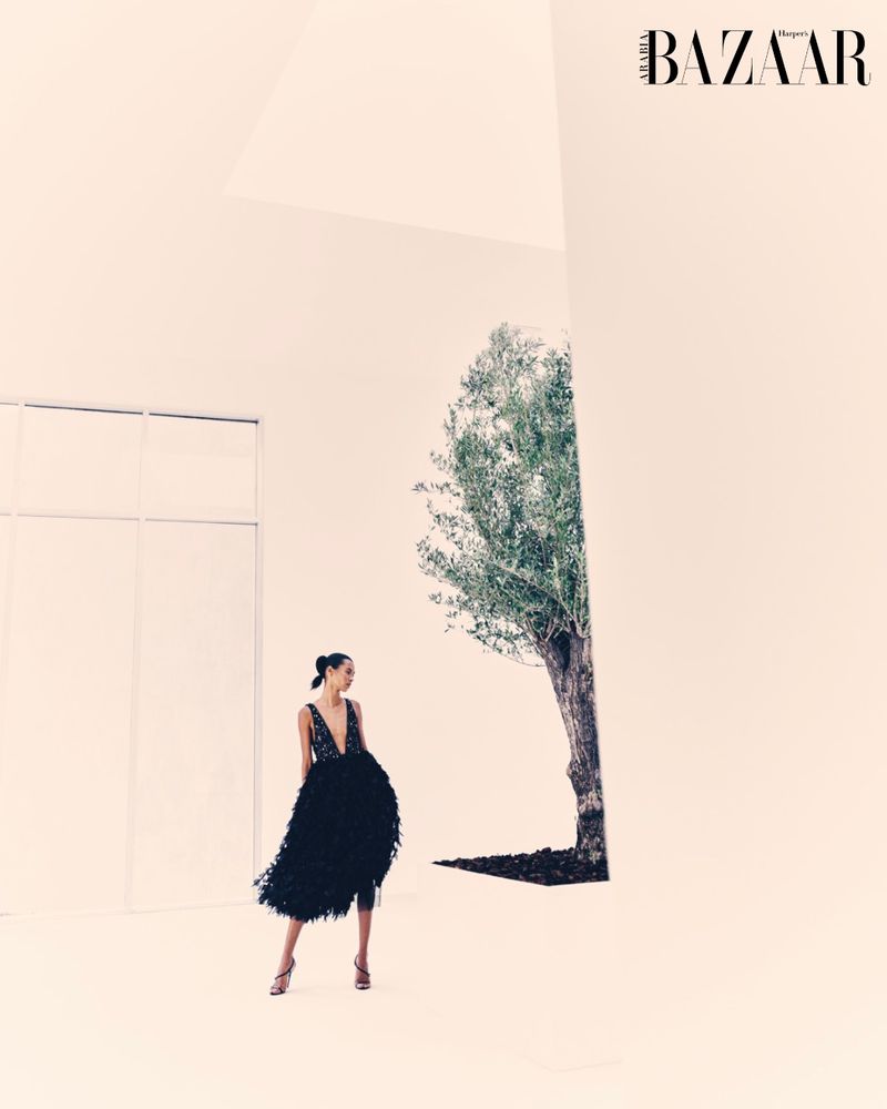 Fashion editorial shot of a woman in a black gown posing next to a tree against a neutral wall, with the Bazaar logo visible.