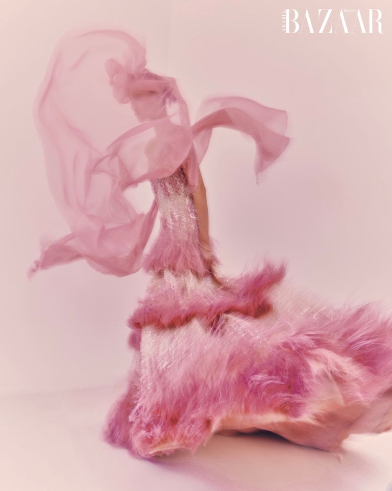 Fashion editorial shot: person in a pink feathered dress with a flowing veil, posing in a studio against a light pink background.
