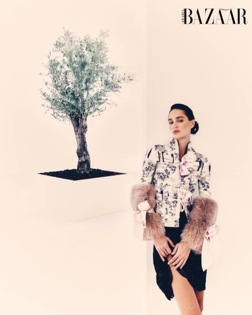 Editorial fashion shot of a woman wearing a floral jacket with fur sleeves and a black skirt, posing near a potted olive tree.