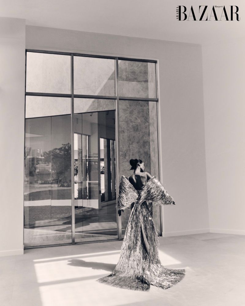 Back view of a woman in a shimmering gown standing in a white room with large windows reflecting the outside environment.