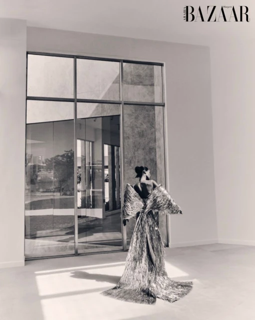 Back view of a woman in a shimmering gown standing in a white room with large windows reflecting the outside environment.