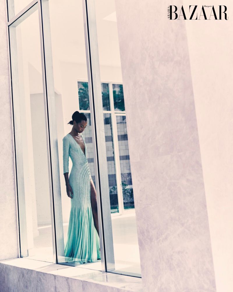 Fashion editorial shot of a Black woman wearing a light blue gown with a thigh slit standing indoors with architectural windows.