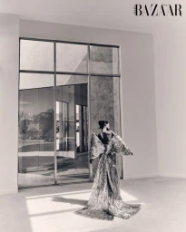 Model standing with back to camera wearing a dramatic fringe gown in front of large glass windows for Harper's Bazaar Arabia