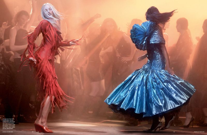 Two women in red and blue couture gowns dancing on a runway in a fashion show with an audience and soft, diffused lighting.