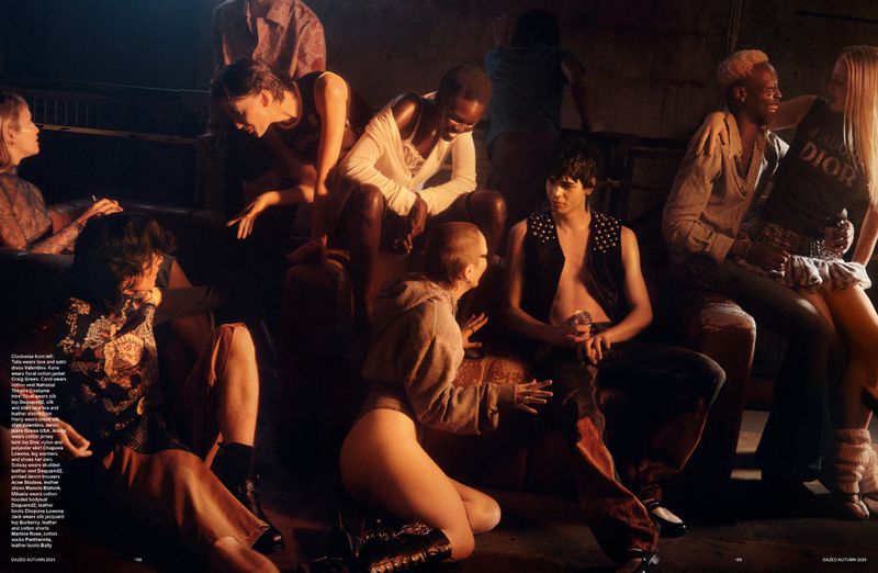 Group of young adults in fashionable clothing gathered together indoors, posing for a fashion editorial shot with warm, moody lighting.