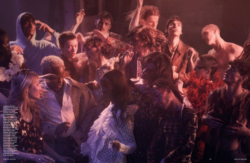Fashion editorial shot of a large group of men and women in a warmly lit indoor space wearing a mix of casual and formal clothing. 