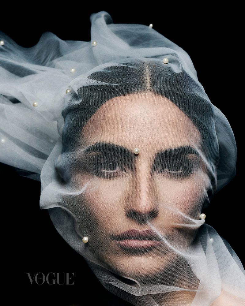 Close-up portrait of a woman with dark hair, wearing a sheer veil adorned with pearls, against a stark black background, in Vogue magazine.