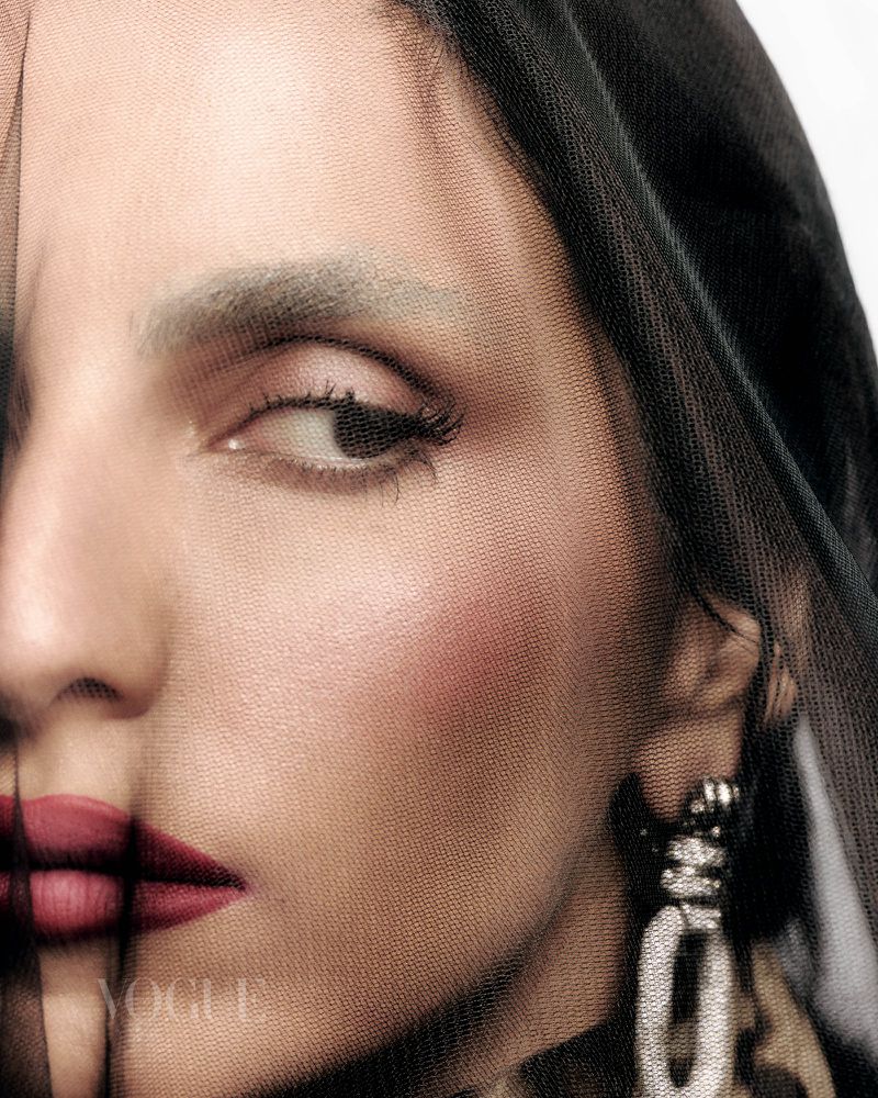 Close-up portrait of an adult female with red lips and fair skin, wearing a black mesh veil and dangling earrings. Vogue logo visible.