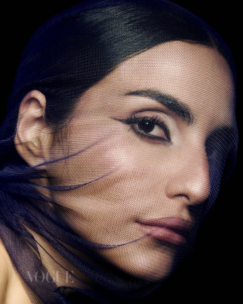 Close-up portrait of a woman with dramatic eye makeup, partially veiled in a dark blue mesh against a stark black backdrop.