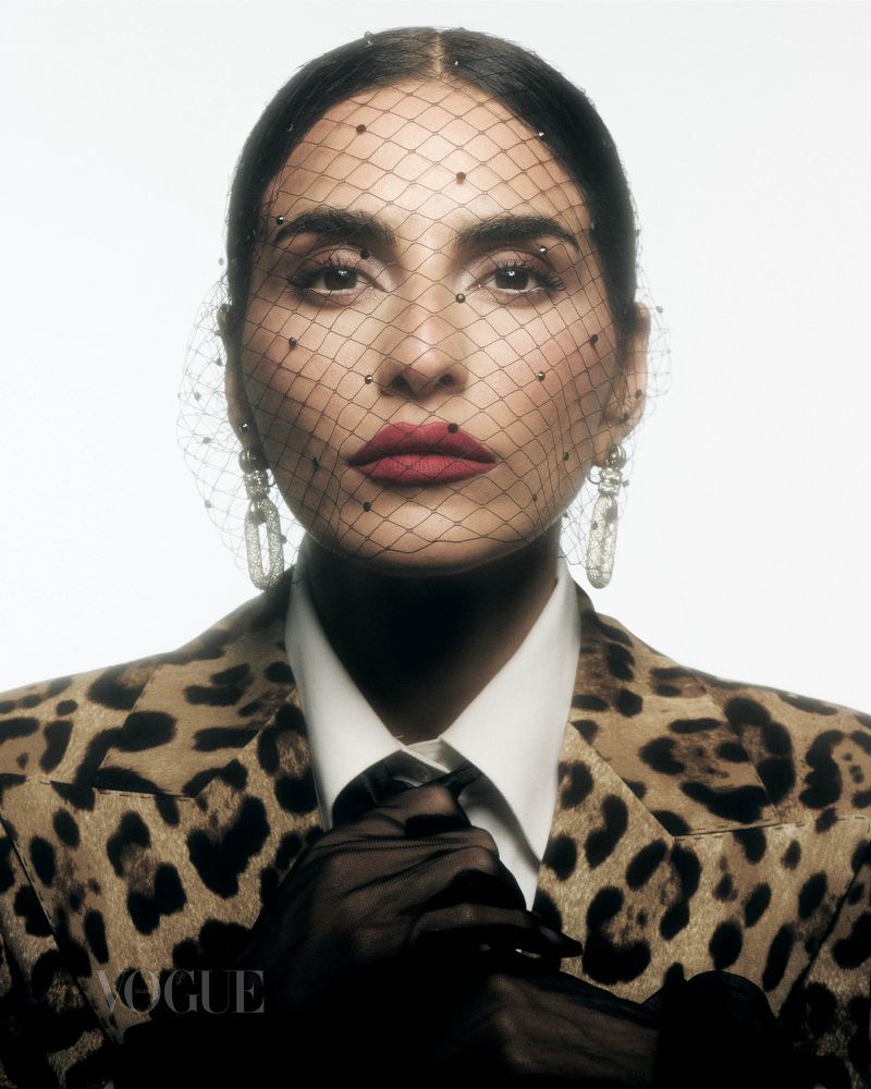 Close-up fashion portrait of an adult female with red lipstick, wearing a leopard print jacket, mesh veil, and statement earrings against white.