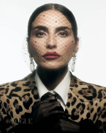 Close-up fashion portrait of an adult female with red lipstick, wearing a leopard print jacket, mesh veil, and statement earrings against white.