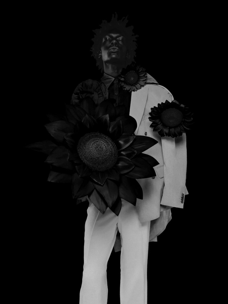 Black and white fashion editorial portrait of a male model wearing a light suit adorned with large dark floral motifs against a black background.
