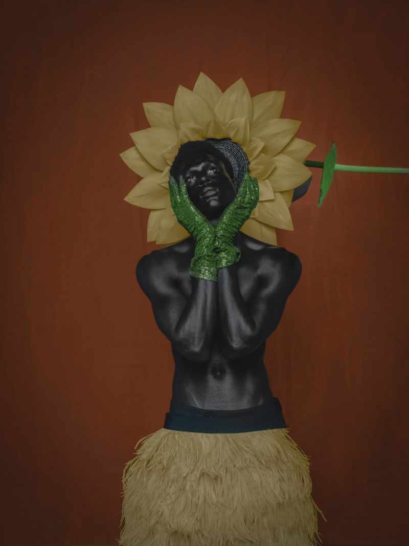 Artistic portrait of a person with dark skin, wearing green gloves and a raffia skirt against an orange studio backdrop.