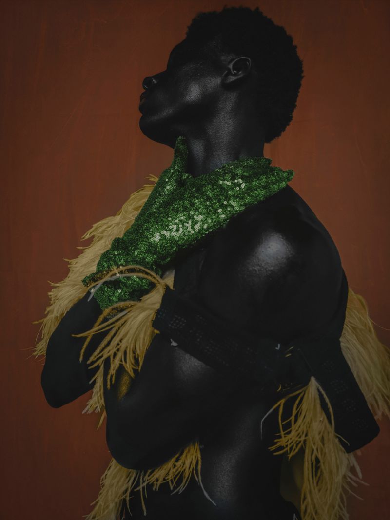 Striking portrait of a shirtless Black male model with green beaded gloves and feather boa against a warm brown backdrop.