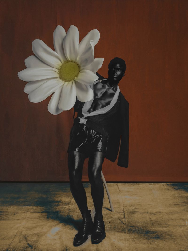 Artistic studio shot of a black man with dark skin, wearing a black jacket and shorts, posing with a large white daisy flower.