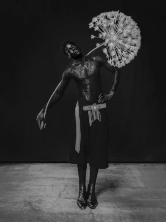 Artistic black and white portrait of a shirtless African American man in a skirt holding a large dandelion against a dark background.