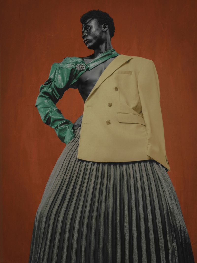 Person posing with mixed textiles in a studio: green satin sleeve, tan blazer, and pleated skirt against a copper background.