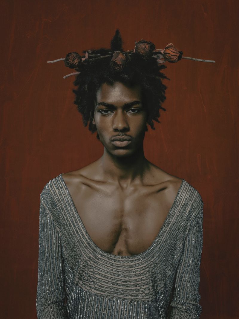 Portrait of a young adult Black male wearing a silver beaded top and a dried floral crown against a mottled rust-red backdrop.
