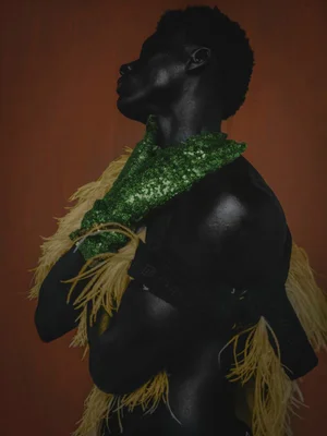 Profile of Black man with glossy skin wearing green sequin gloves and feather trim for Numero Netherlands