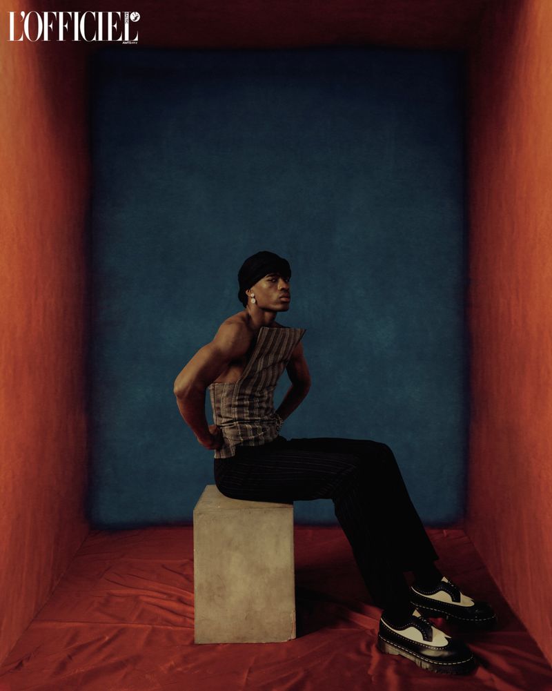 Man poses seated in a square photo studio wearing a sleeveless striped top, dark pants, and black and white Oxford shoes.