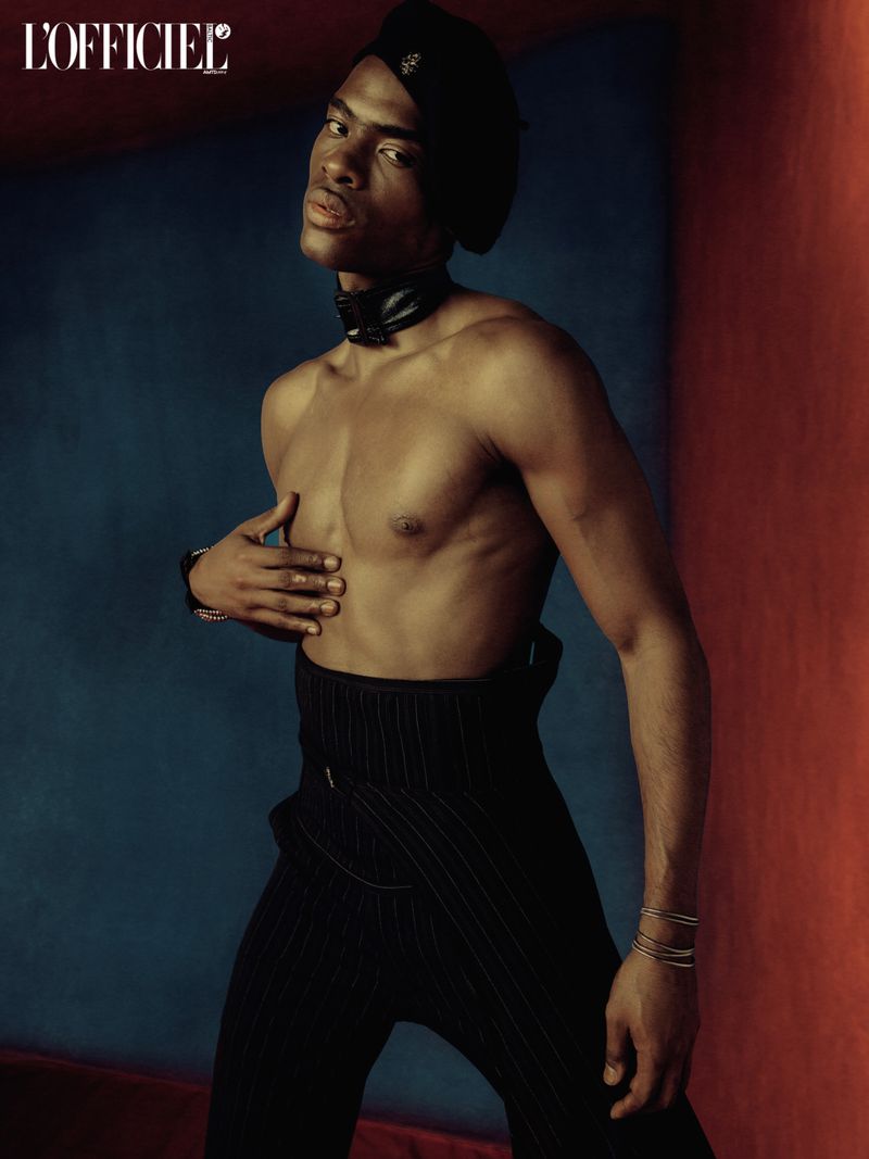 Shirtless African American man poses against a split blue and red background wearing black pants, bracelets, and a matching newsboy cap.