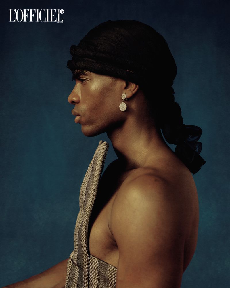Portrait of a dark-skinned adult male model with head wrap and earring, wearing a structured garment in front of a blue background.