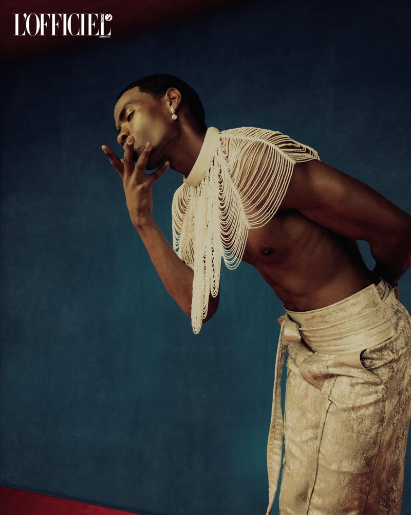Portrait of an adult Black male model with beaded garment, wearing tan pants, set against a blue backdrop. Fashion editorial.