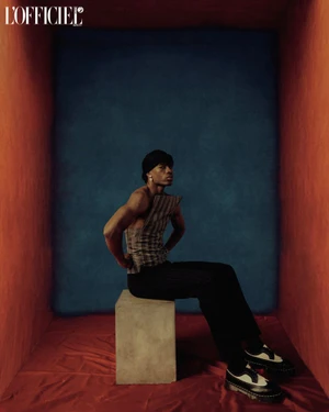 A man wearing a structured plaid top and black trousers seated on a cube inside a red and blue box-like set.