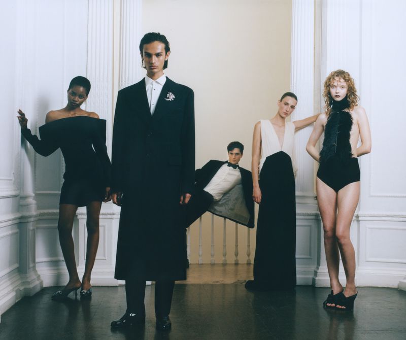A group of five fashion models pose in formal wear indoors. Two women wear dresses and two men wear tuxedos. One woman wears a bodysuit.