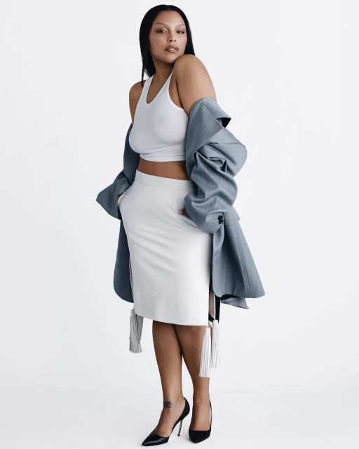 A woman poses in a white pencil skirt, white tank top, and a gray blazer in a full shot in a photo studio setting.