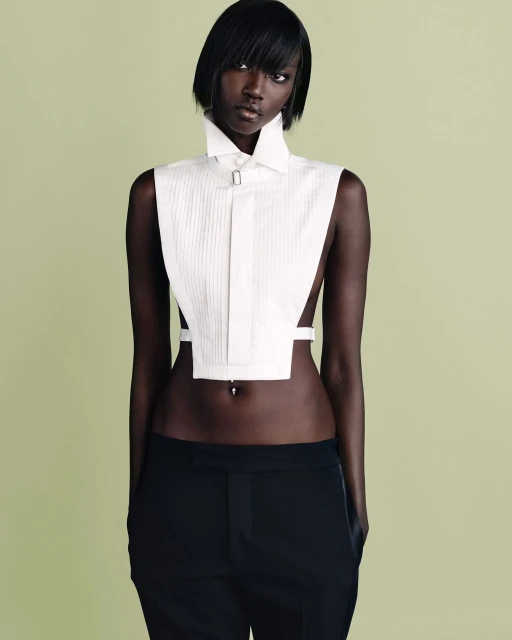 A slim, dark-skinned woman with black hair poses in an open collared white top and black pants against a light green backdrop.