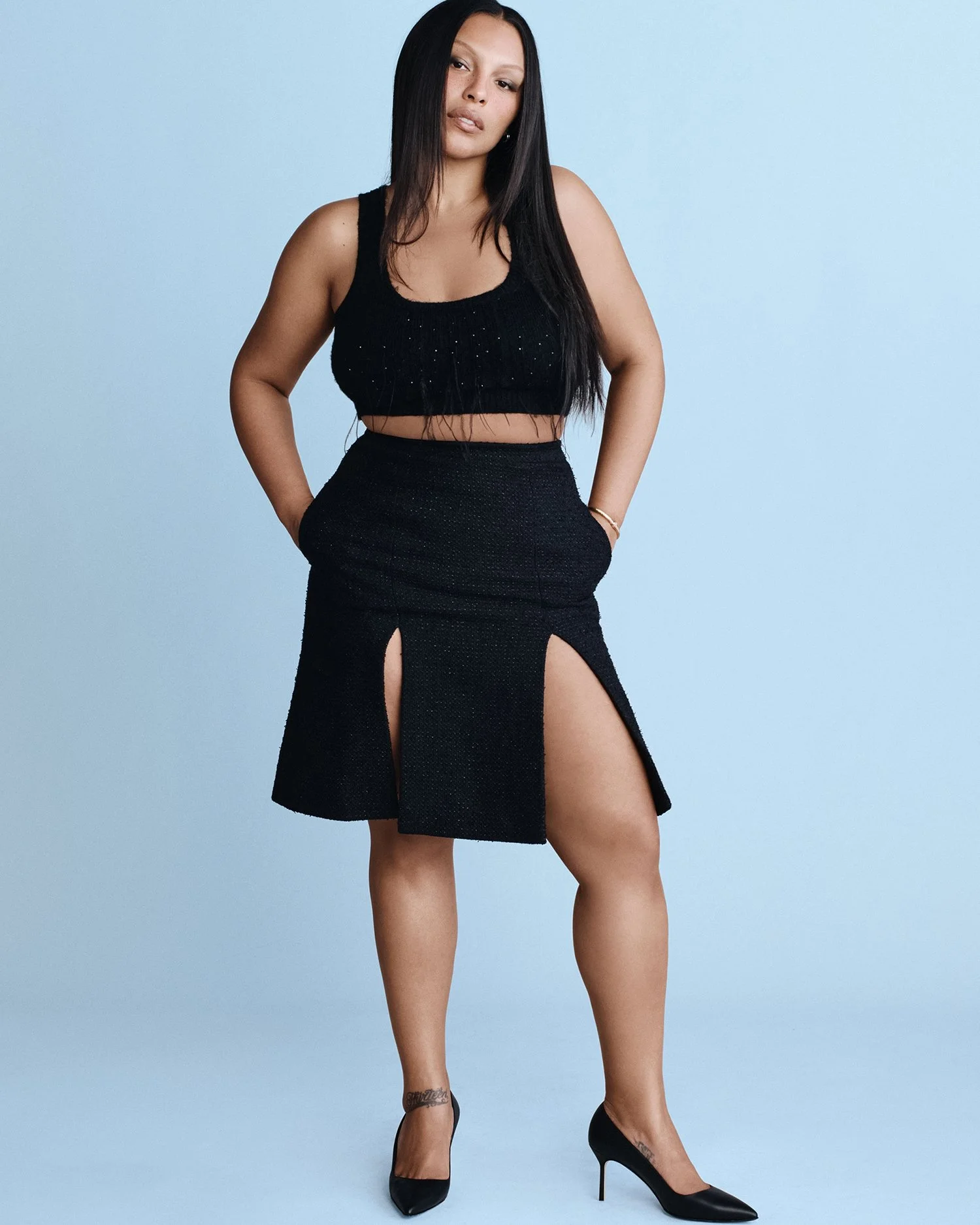 Full-body portrait of a woman in a black crop top and skirt, hands in her pockets, posing against a light blue background.