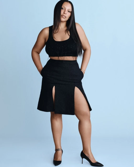 Full-body portrait of a woman in a black crop top and skirt, hands in her pockets, posing against a light blue background.
