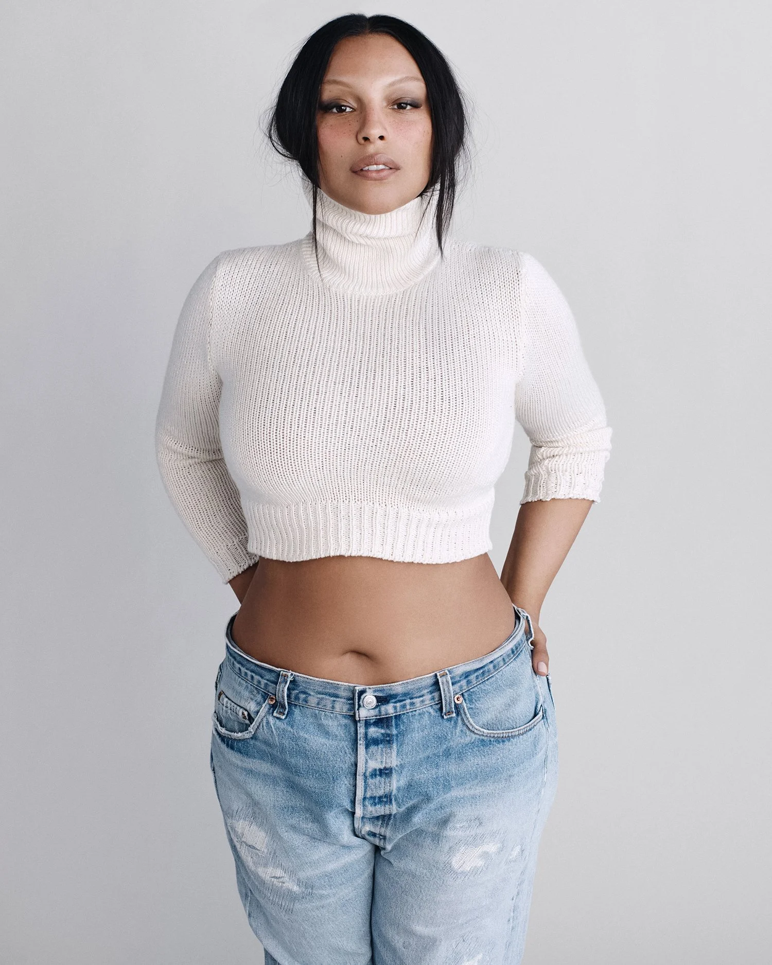 Paloma Elsesser in a cropped white turtleneck sweater and light blue jeans against a neutral studio background