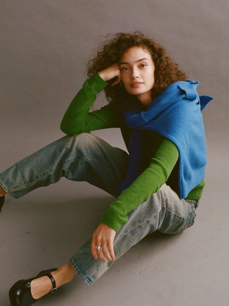 A young woman with curly hair poses indoors on a gray floor wearing a green sweater, blue vest, jeans, and black shoes in a studio shot.