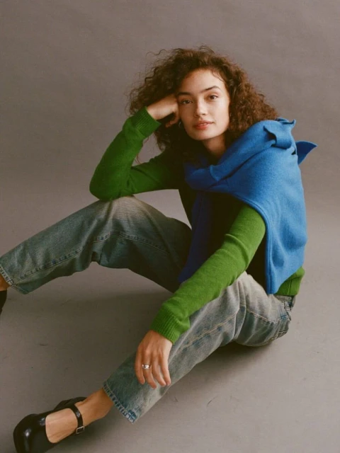 A young woman with curly hair poses indoors on a gray floor wearing a green sweater, blue vest, jeans, and black shoes in a studio shot.