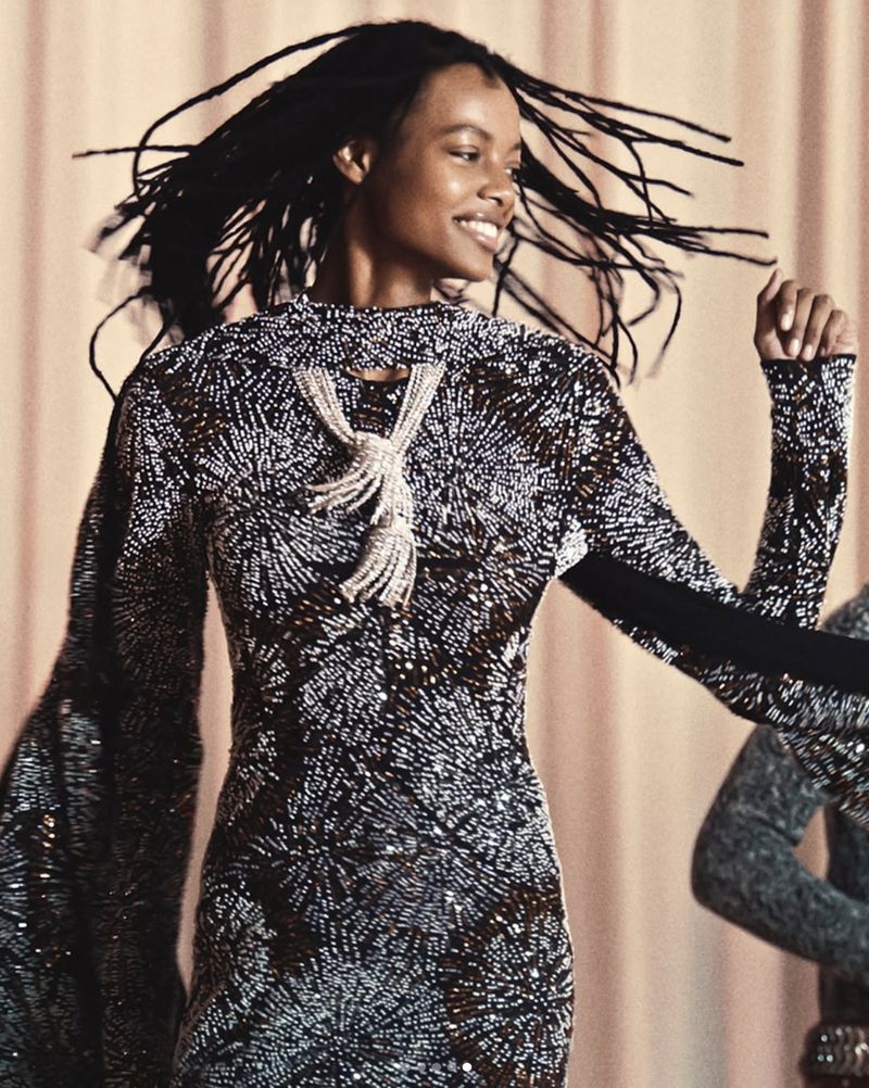 A smiling woman with long dark hair is dancing while wearing an elaborately beaded, long-sleeved dress with a draped beaded necklace.