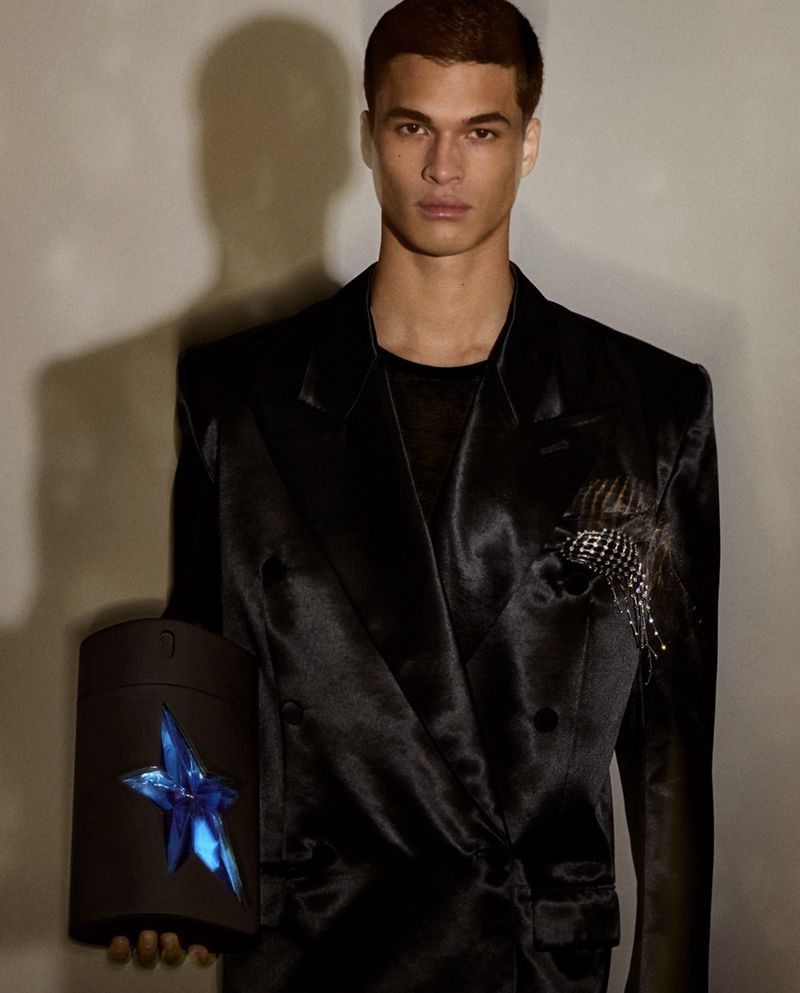 A stylish adult male poses indoors wearing a black blazer. The man is holding a black container with a blue star on the side.