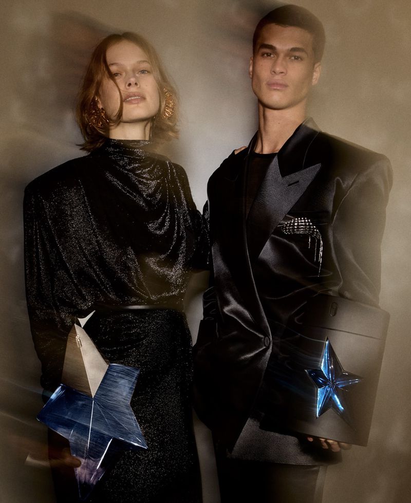 Elegant fashion shot of a woman and man wearing black outfits with star accents, posing indoors with soft lighting, showcasing modern style.