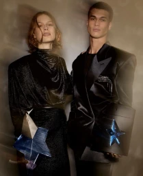 Two fashion models in black clothing holding star-shaped perfume bottles against a smoky abstract background.