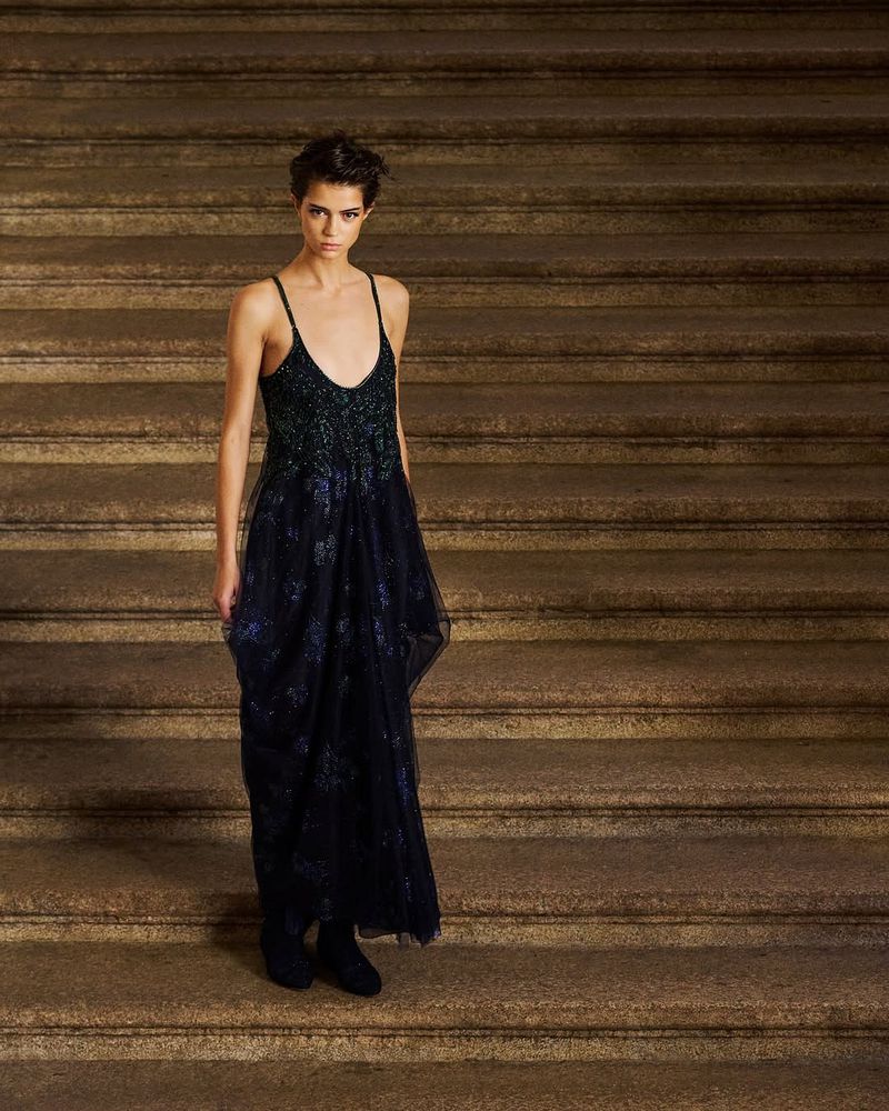 A fair-skinned fashion model poses on stone steps wearing a sleeveless black dress with embroidered floral accents in a medium shot.