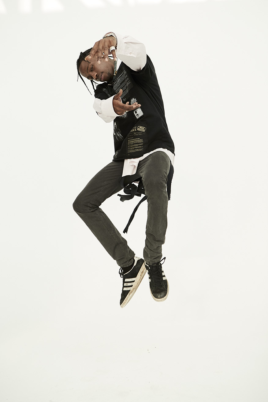 Man mid-air, stylishly dressed in black top and grey pants, poses with hands near his face against a stark white backdrop.